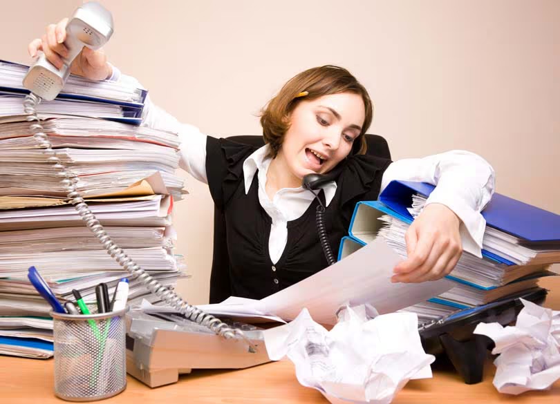 書類と電話に囲まれ、仕事に追われる忙しい社会人女性の様子